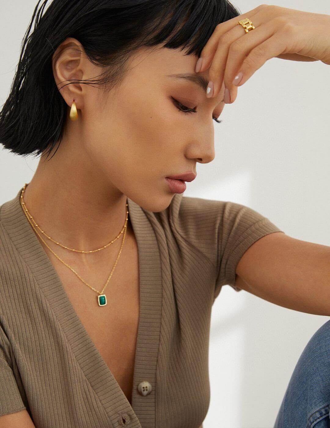 Woman wearing gold jewelry including earrings, necklace, and ring against a neutral background