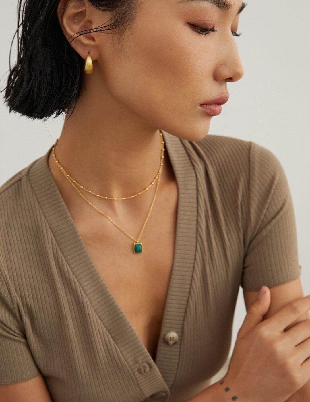 Woman wearing a gold necklace with a green gemstone, looking away from the camera.