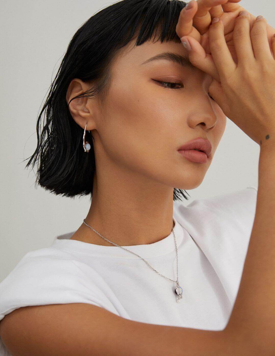 Woman with short black hair wearing a white top and silver jewelry against a plain background