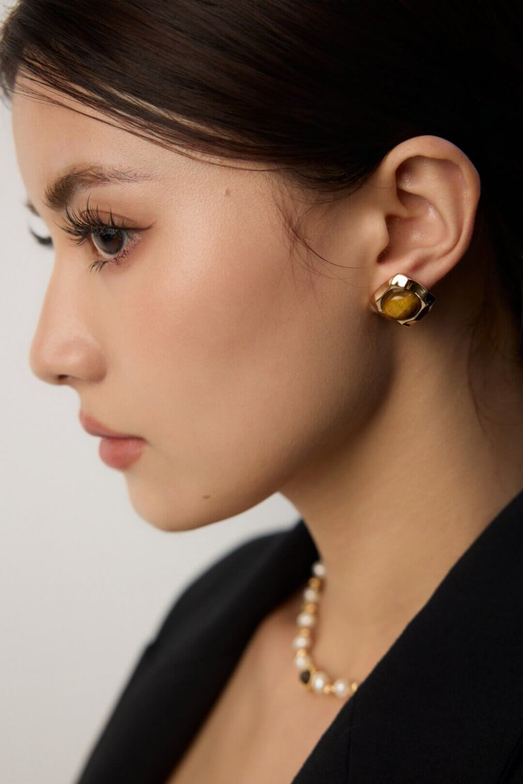 Close-up of a person wearing a yellow gemstone earring and pearl necklace against a neutral background