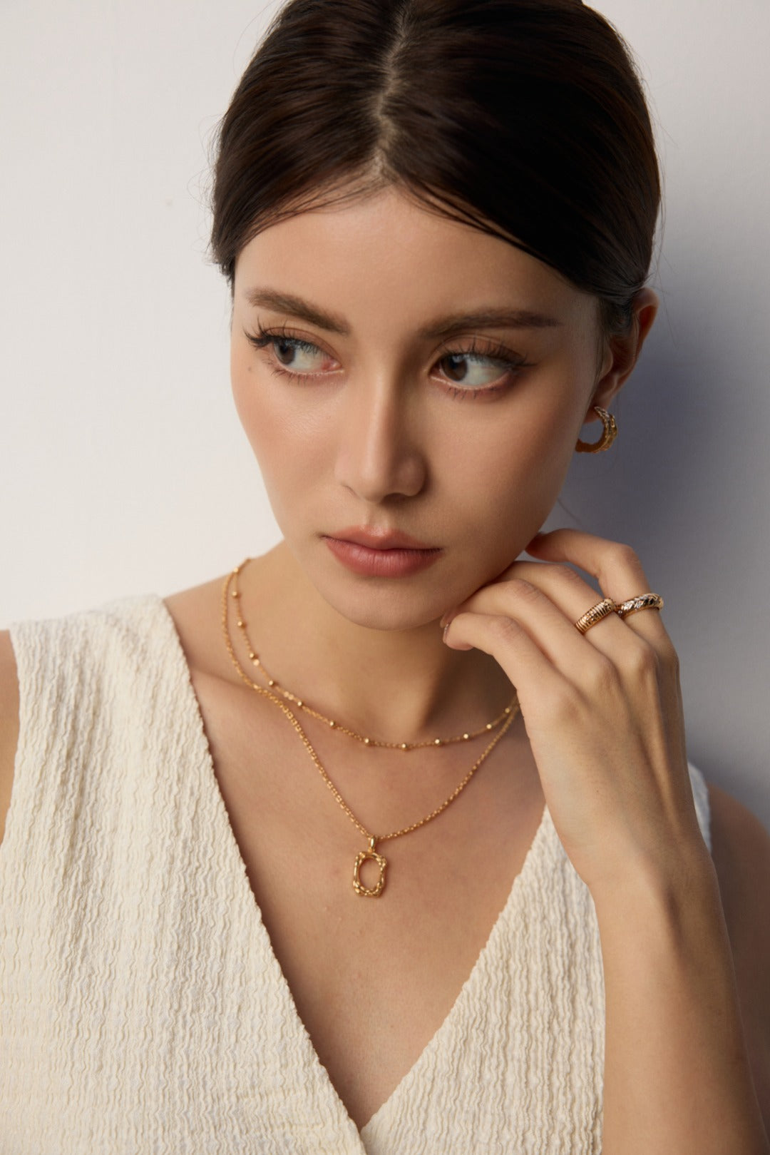 Woman wearing gold jewelry including a necklace, ring, and earrings against a neutral background