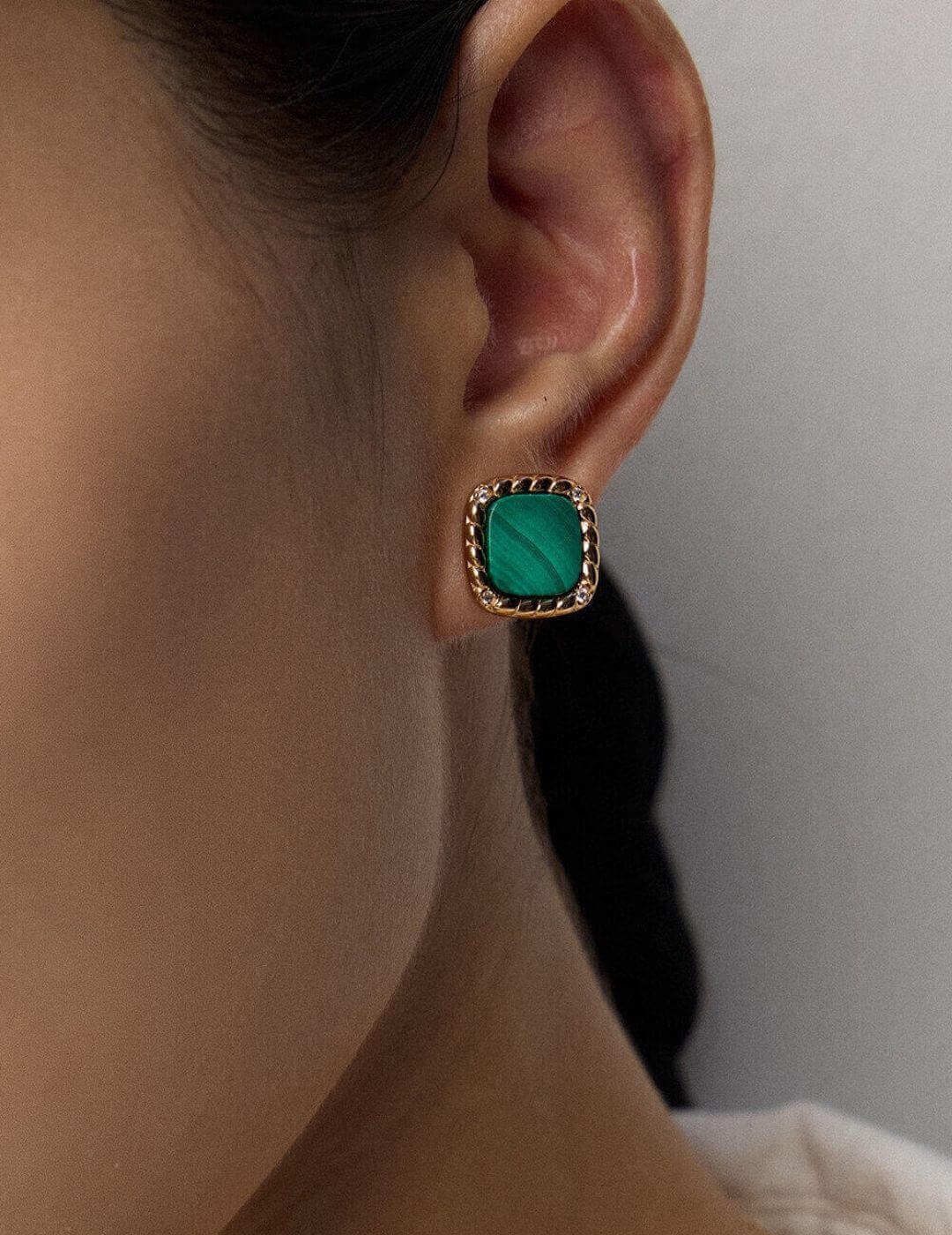 Close-up of an ear wearing a green earring with gold frame against a neutral background